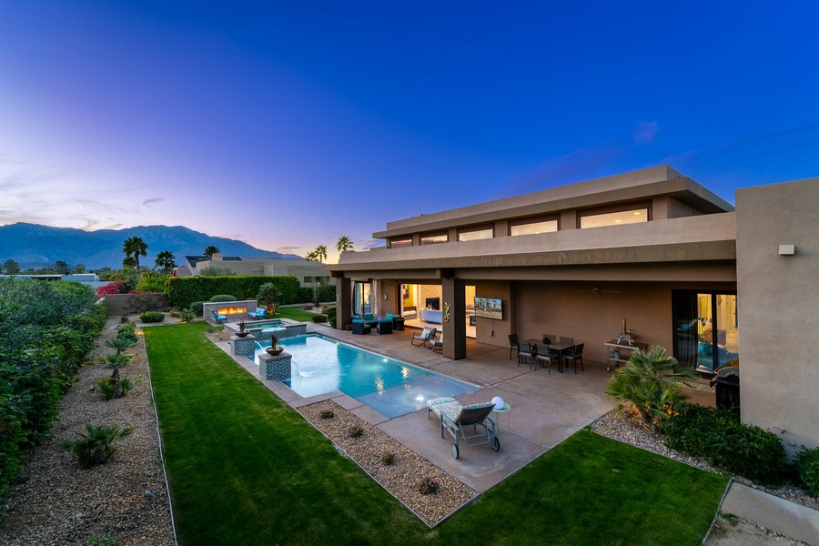 Glass-walled home and lit pool at dusk, mountains behind
